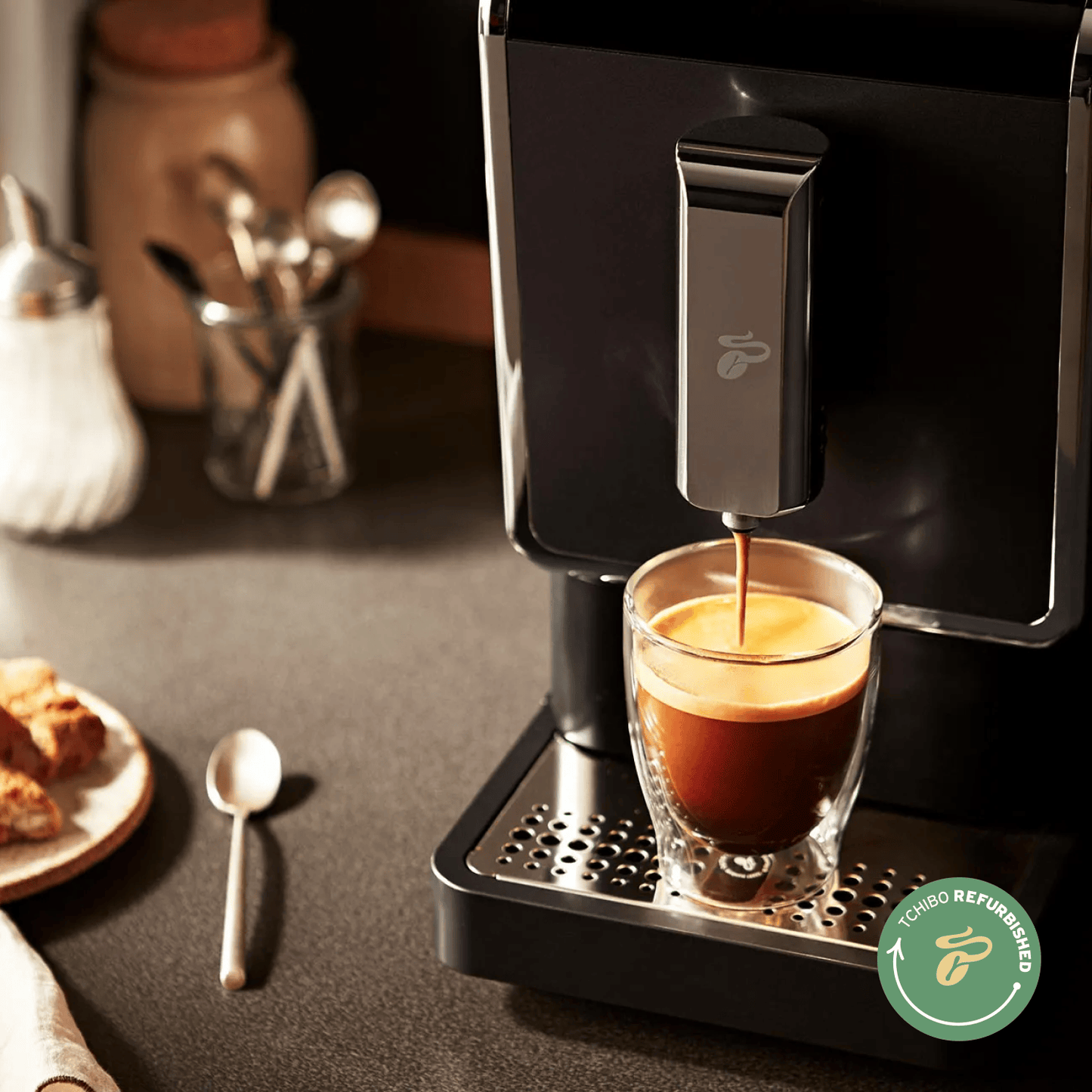 Nahaufnahme des Tchibo Esperto Caffè in Anthracite der gerade eine Tasse Kaffee brüht. Neben der Maschine sind weitere Küchenutensilien erkennbar.