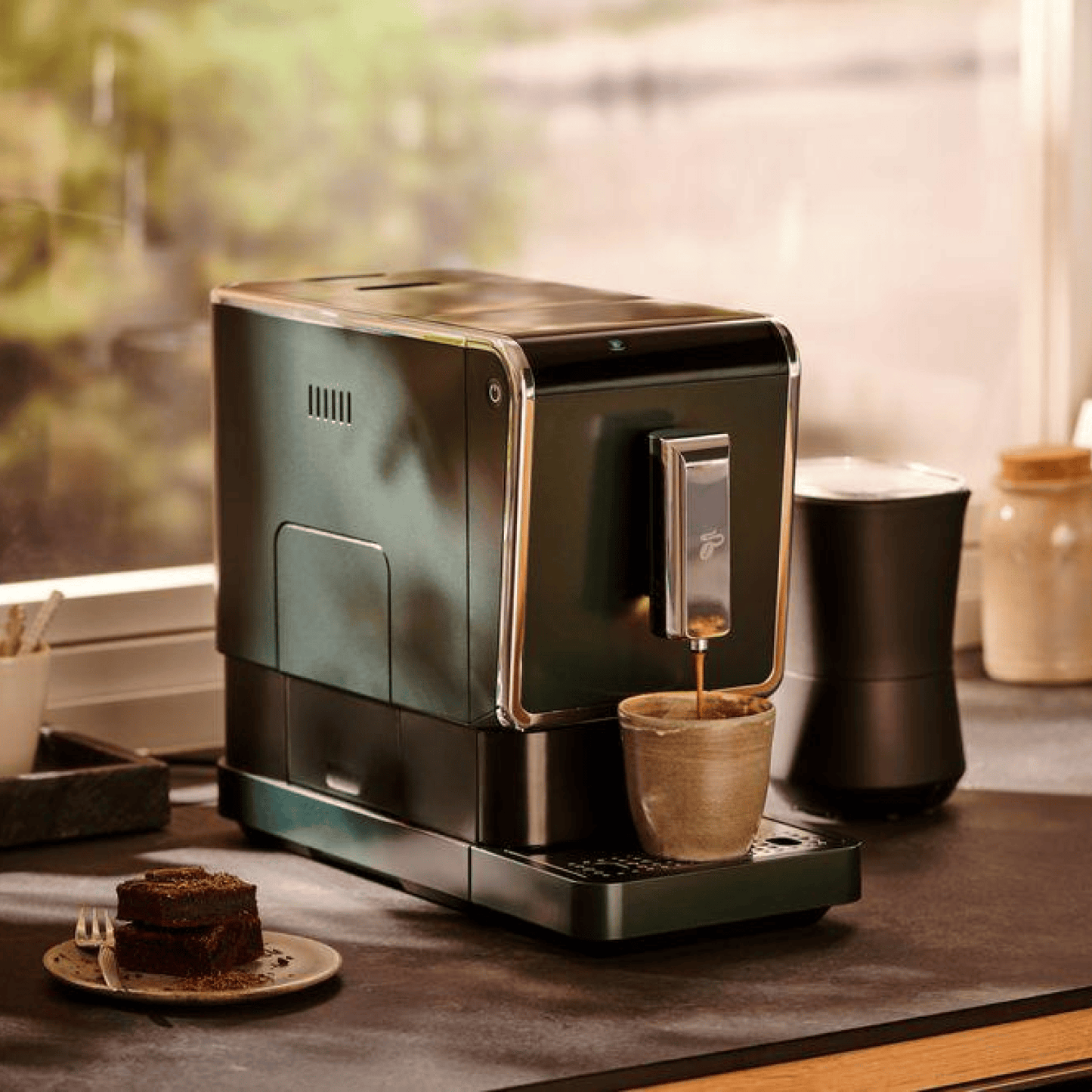 Der Tchibo Esperto Caffè in Dark Green steht auf einer Anrichte, während gerade eine Tasse Kaffee gebrüht wird. Ein Kuchen ist neben der Maschinen zu sehen.