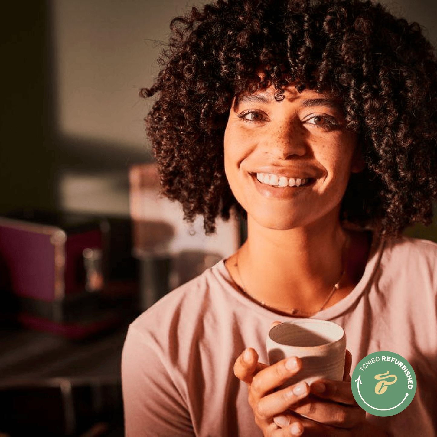 Eine Frau mit Sommersprossen lächelt in die Kamera und hält zufrieden eine Tasse Kaffee in beiden Händen, die mit dem Tchibo Esperto Caffè gebrüht wurde.