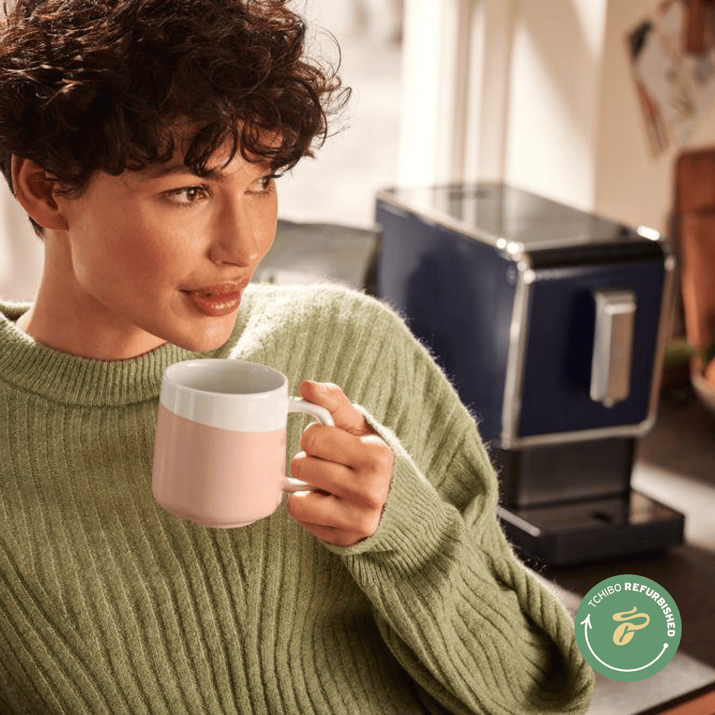 Eine Frau mit kurzen lockigen Haaren hält eine Tasse Kaffee in der Hand. Im Hintergrund ist eine Tchibo Esperto Caffè in Night Blue sichtbar.