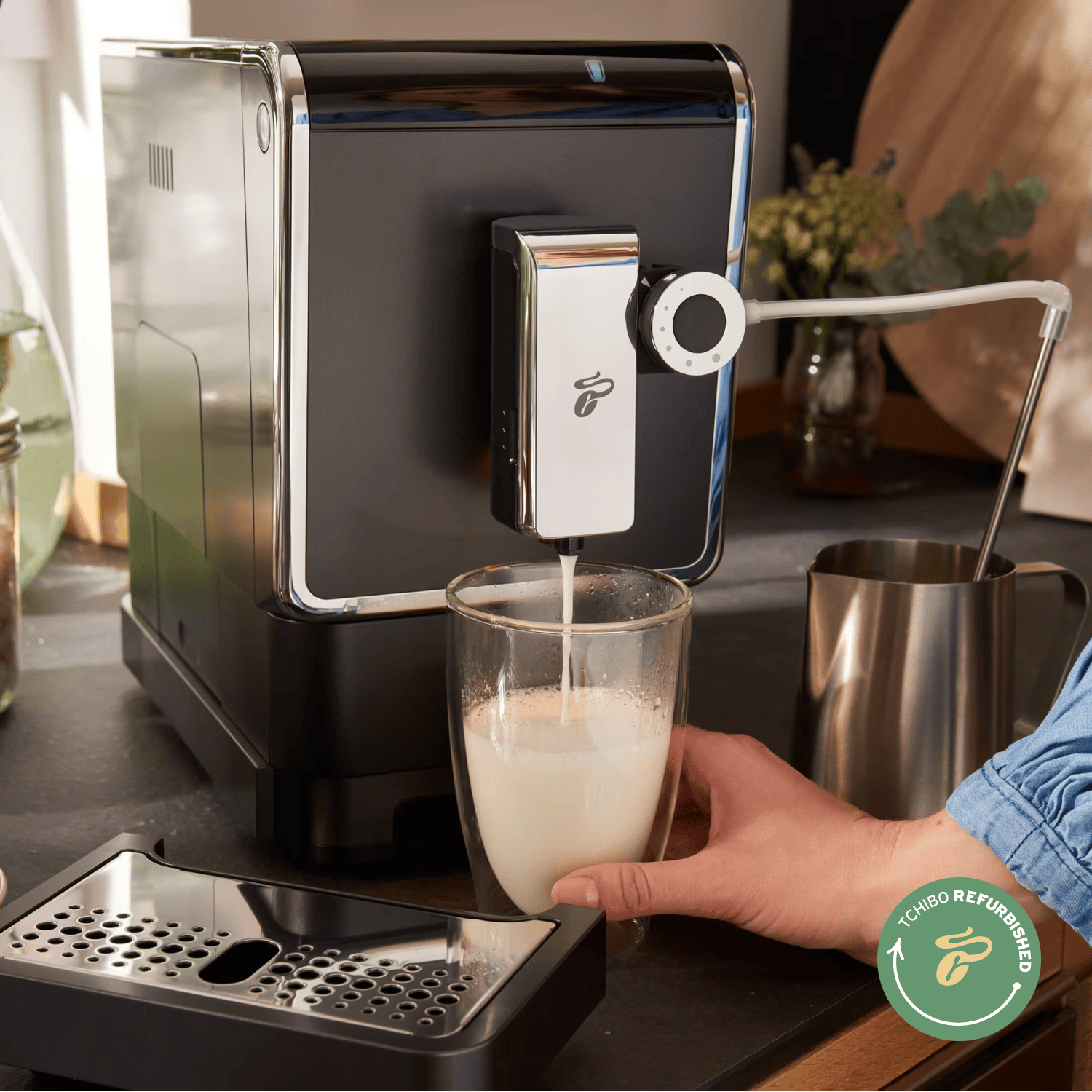 Tchibo Esperto Pro die Milchschaum im Glas zubereitet, eine Hand hält das Glas fest und man sieht das "Tchibo Refurbished"-Logo