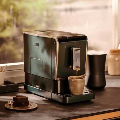 Der Tchibo Esperto Caffè in Dark Green steht auf einer Anrichte, während gerade eine Tasse Kaffee gebrüht wird. Ein Kuchen ist neben der Maschinen zu sehen.