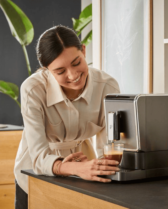 Frau steht an einer Anrichte und brüht sich eine Tasse Kaffee mit dem Tchibo Esperto Caffè in Silver. Sie hält die Tasse am Auslauf fest und kann den Kaffee kaum erwarten.