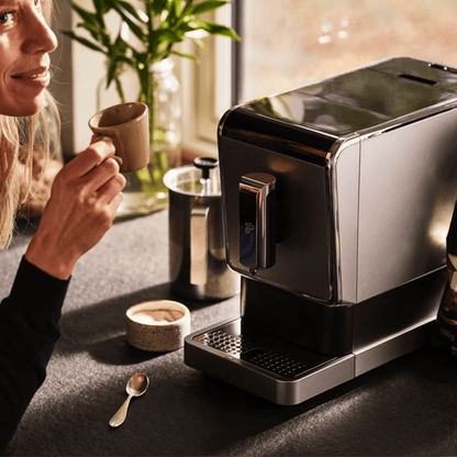 Der Tchibo Esperto Caffè in Silver steht auf einer Küchenanrichte. Am Rand des Bildes ist eine Frau erkennbar, die gerade aus einer Tasse Kaffee trinkt. Auf der Anrichte sind weitere Gegenstände wie ein Löffel und ein Milchaufschäumer sichtbar.