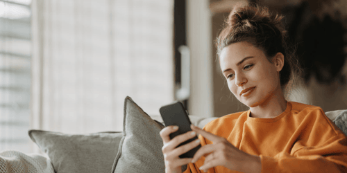 Frau sitzt auf einem Sofa, lächelt und schaut auf ihr Handy, während sie den Tchibo Help Service durchsucht, um Unterstützung oder Informationen zu finden.