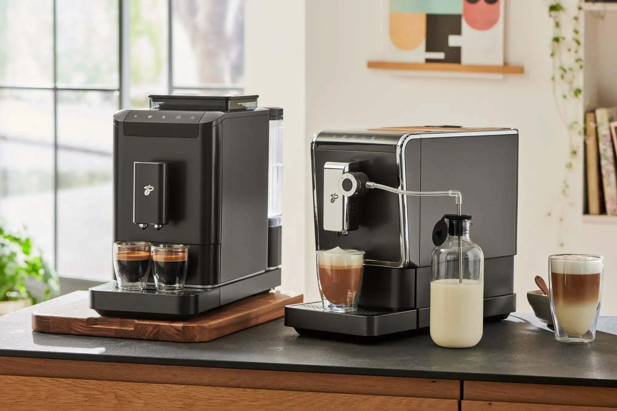 Zwei schwarze Tchibo-Kaffeevollautomaten auf einer Küchentheke bereiten Espresso und Milchschaum zu.