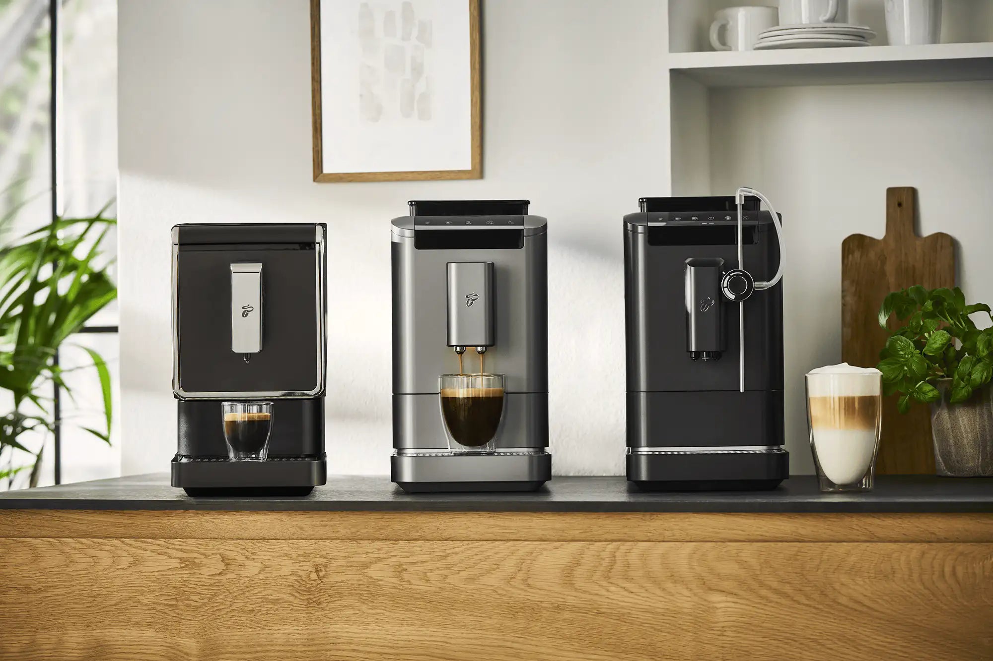 Drei Tchibo-Kaffeevollautomaten auf Holztisch mit Espresso, Cappuccino und Milchschaum in Gläsern sowie Pflanzenkübeln im modernen Küchenraum.