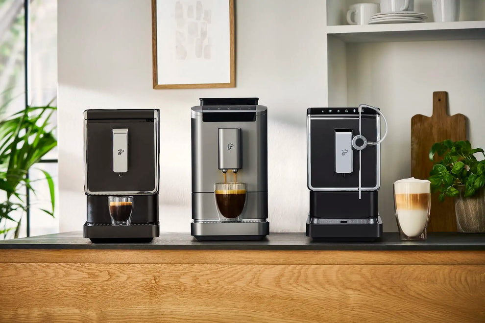 Drei Tchibo-Kaffeevollautomaten auf Holztisch mit Espresso, Cappuccino und Milchschaum in Gläsern sowie Pflanzenkübeln im modernen Küchenraum.