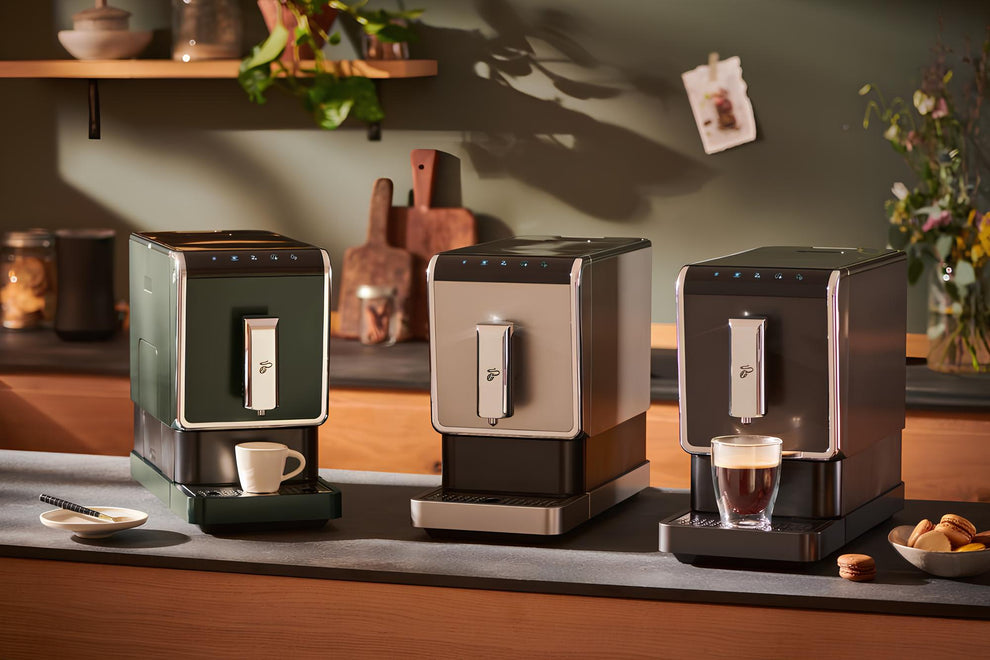 Drei Tchibo Esperto Caffè Kaffeevollautomaten der ersten Generation in verschiedenen Farben nebeneinander auf einer Küchenanrichte. Vor den Maschinen liegt französisches Gebäck, während die rechte Maschine gerade eine frische Tasse Kaffee gebrüht hat – dampfend und servierbereit.